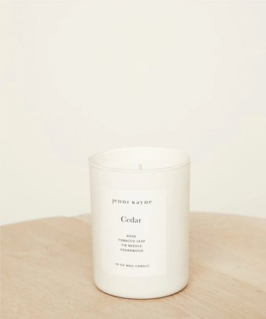 Cedar Glass Candle from Jenni Kayne on a wood table. The vessel is matte white glass with a label that says "Jenni Kayne" and "Cedar," along with the scent notes.