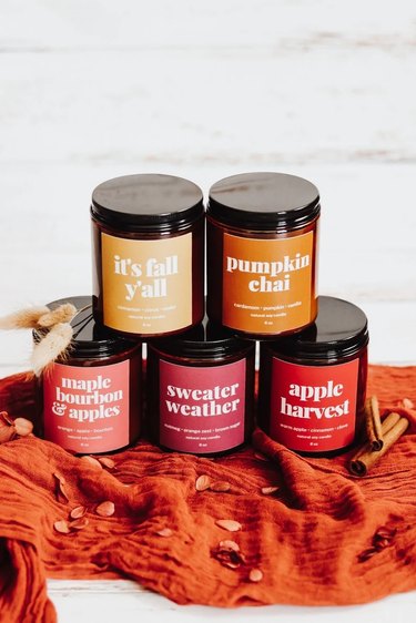 Five fall candles from Etsy on a piece of orange linen fabric. The candles come in dark brown glass jars and have eye-catching labels in bold fonts.