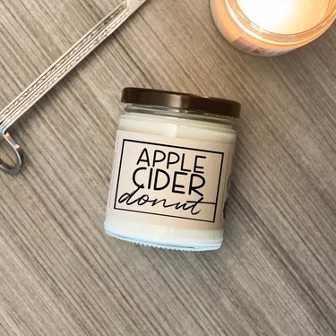 Apple Cider Donut Candle from Etsy on a wooden background. It comes in a clear glass jar and has a bronze-colored metal lid.