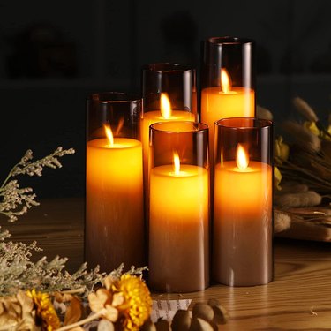 Flickering Flameless Candles from Amazon in dark brown glass hurricanes on a wooden table surrounded by dried foliage.