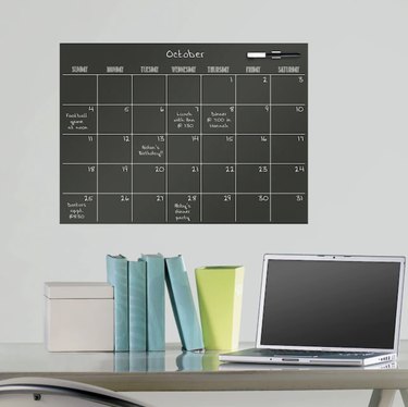 Black wall calendar hanging over desk with computer. The writing on it is white and little notes are scribbled on the calendar.