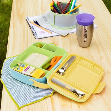 Green, blue, and yellow Munchkin Toddler Bento Box on a wooden table outdoors. The bento box includes a stainless steel spoon and spork.