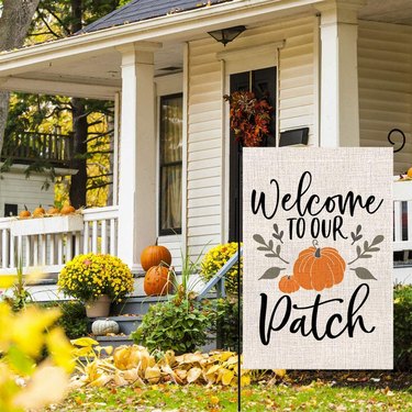 Welcome Pumpkin Patch Burlap Yard Flag from Amazon on a flag stand in front of a house. The flag says 'Welcome to our patch' and has an image of a pumpkin.