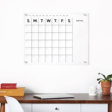 Acrylic wall calendar hanging over desk. The days of the week are labeled by their first letter and there's a section for writing notes.