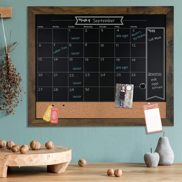 Black chalk calendar inside rustic brown frame with notes and days written in chalk pen.