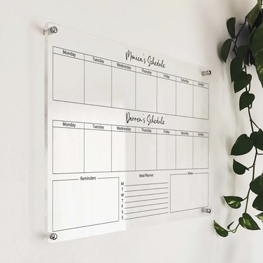 Acrylic wall calendar hanging on a white wall. The top says "Monica's Schedule" and the bottom says "Darren's Schedule."