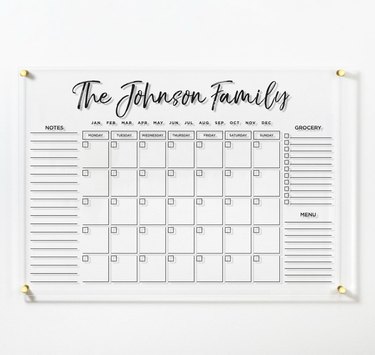 Acrylic, personalized white wall calendar hanging on wall. The calendar says "The Johnson Family" in cursive and has gold hardware.