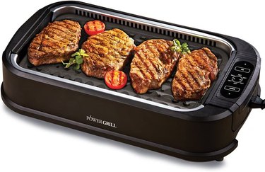 Power XL indoor smokeless grill, shown on a white ground with four well-seared steaks on the cooking surface, garnished with grilled tomatoes and fresh herbs