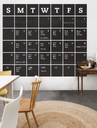 Chalkboard-colored wall calendar that takes up an entire wall. The days of the week are marked by their first letter and notes are written on it in chalk pen.