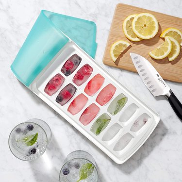 OXO no-spill ice cube tray shown in an overhead view, containing ice cubes with berries and citrus frozen into them, flanked by glasses with ice and a cutting board with knife and lemon slices