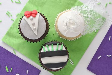 Three cupcakes: one shaped like a tuxedo with red bow tie, one designed like a bridal veil with a ring on top and one with black stripes and smaller green vertical stripes