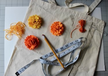 Felt marigolds, a measuring tape, a pencil and thread atop a plain canvas tote bag