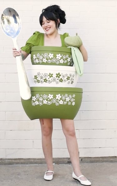 Woman holding a large silver spoon while dressed up as a stack of green and white vintage Pyrex mixing bowls with white and green flowers