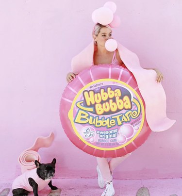 Woman dressed up in pink bubble gum tape costume next to a dog wearing a pink bubble gum costume