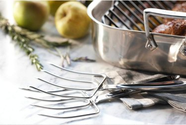All-Clad turkey forks on a white countertop next to a roasting pan with a turkey in it.