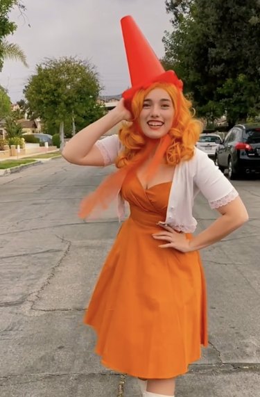 Woman dressed in an orange dress and white sweater with an orange traffic cone on her head