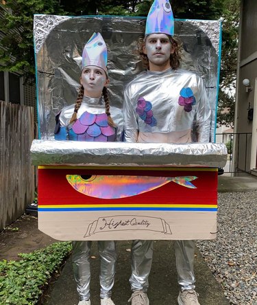 Two people dressed in silver clothing standing inside a box shaped like a sardine tin