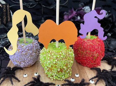 A purple, bright green and red caramel apple decorated with sprinkles and cutouts of hairstyles