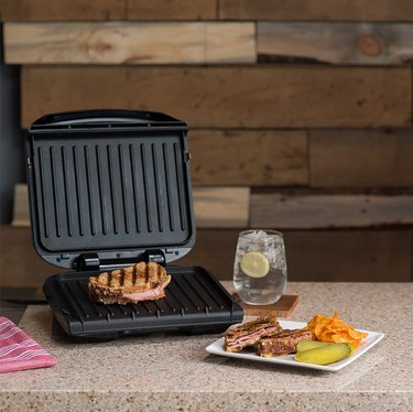 George Foreman grill and panini press, pictured on a stone countertop in front of rustic wood wall, shown open with a sandwich on the grill, another on a plate in front, and a fizzy beverage to the side