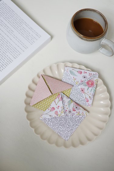 Three origami corner bookmarks made with decorative paper on a plate next to a book and coffee