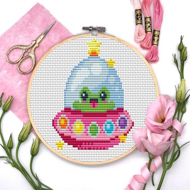 Round cross-stitch pattern with a green frog inside a pink spaceship