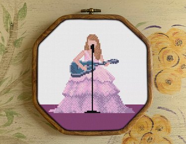 Cross-stitch design featuring Taylor Swift in a pinkish-purple dress holding a blue guitar at a microphone