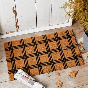 Coir doormat with black plaid lines on a wood floor with a rolled-up newspaper laying on it.