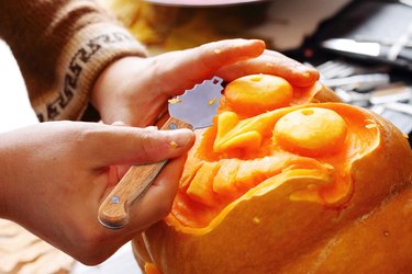 Woman carving an intricate face into a pumpkin with a wooden-handle tool.