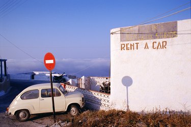 An older style car parked outside of a building with a sign reading ?Rent a Car'; Greece.