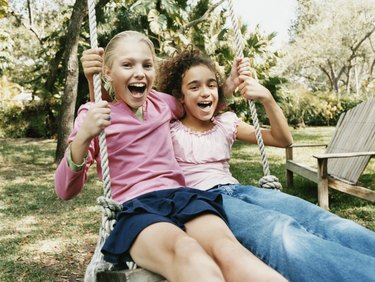 Two Young Girls Sit Side by Side on a Swing in a Garden, Laughing