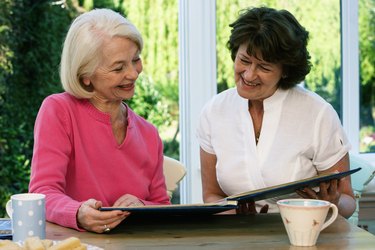 Two senior women sitting at kitchen table looking at album, smiling