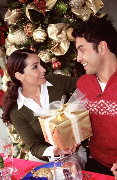 Woman handing man gift at dinner table by christmas tree