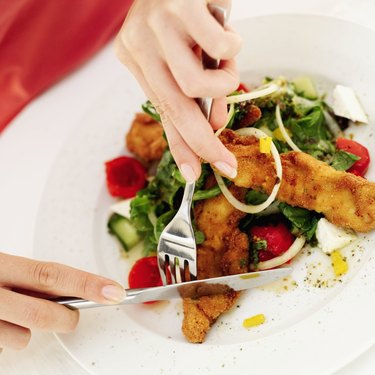 Close-up of a woman's hands slicing a piece of fried fish with fork and knife