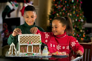 A brother and sister decorate a gingerbread house at Christmastime.