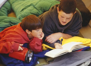 Two brothers (10-14) lying in sleeping bags, reading book in tent