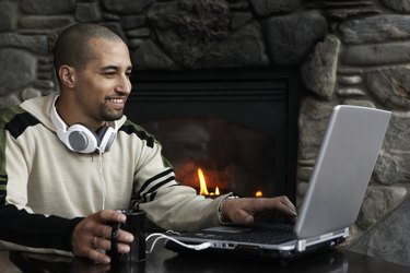 Man with coffee cup using laptop beside fireplace, smiling