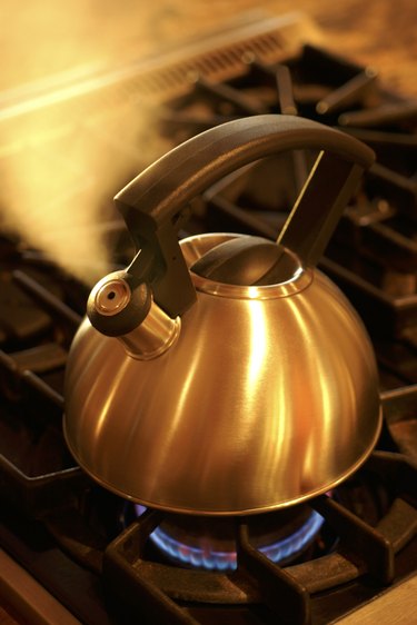 A stainless steel kettle blows steam while sitting on a hot burner of a stove.