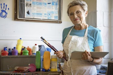 "Mature woman in art classroom, holding brushes and paper, portrait"