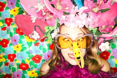 A woman looks cross-eyed at the plastic bug on her nose while wearing a wacky hat of flowers and orange sunglasses in front of a flowered background.