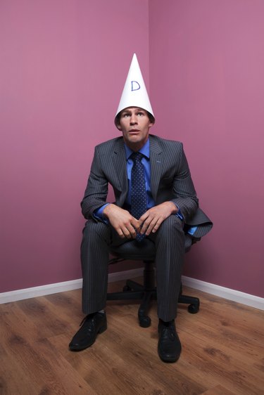 Businessman sat in corner wearing dunce hat