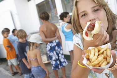 Girl (8-10) eating hotdog, children (6-12) queuing at vendor's window