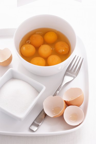 Egg yolks and egg whites in bowl, with eggshells, fork and sugar bowl on tray, close-up