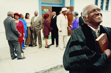 Smiling Priest Standing in Front of a Group of People Outside a Church Holding a Bible