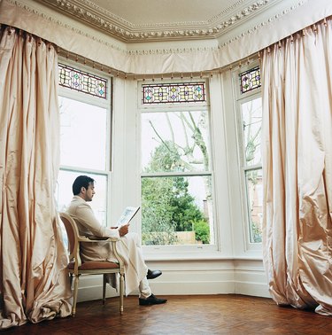 Young Man Sitting in a Chair by a Window at Home Reading a Newspaper