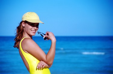 Young woman wearing cap and holding sunglasses on beach, smiling 