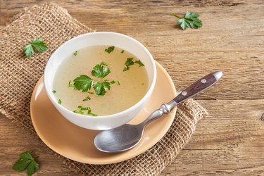 Chicken broth with parsley in white bowl over wooden background with copy space