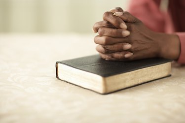 Senior woman sitting at table with hands clasped on Bible, close-up of hands