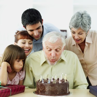 close-up of a senior man blowing candles on a birthday cake with his family standing behind him