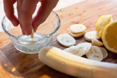 Hand dipping banana slices into a small bowl filled with lemon juice.