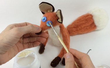 Female hands painting glossy Mod Podge onto the needle felted fox's button eyes.